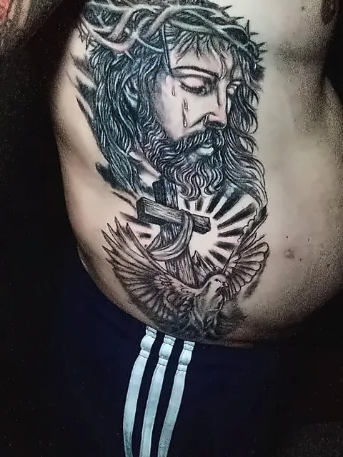 Tatuagem em preto e cinza na lateral do tronco: rosto de Jesus com coroa de espinhos, cruz com raios de luz e uma pomba